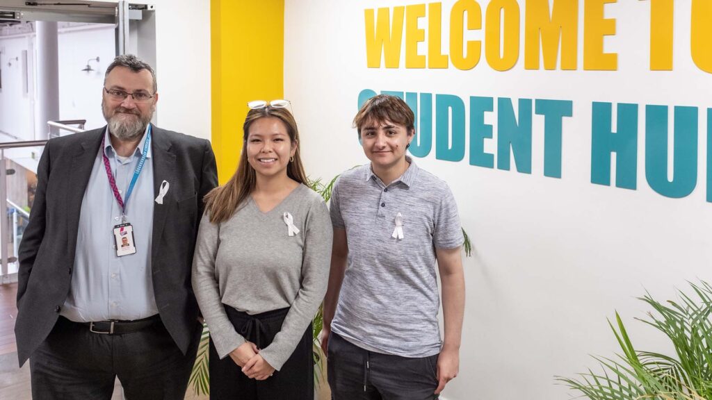 Trevor, Rachel and Atlas stood together inside the Students' Union building wearing white ribbons and looking at the camera