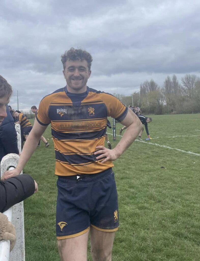 Harry, student recruitment on a rugby pitch smiling with hand on his hip