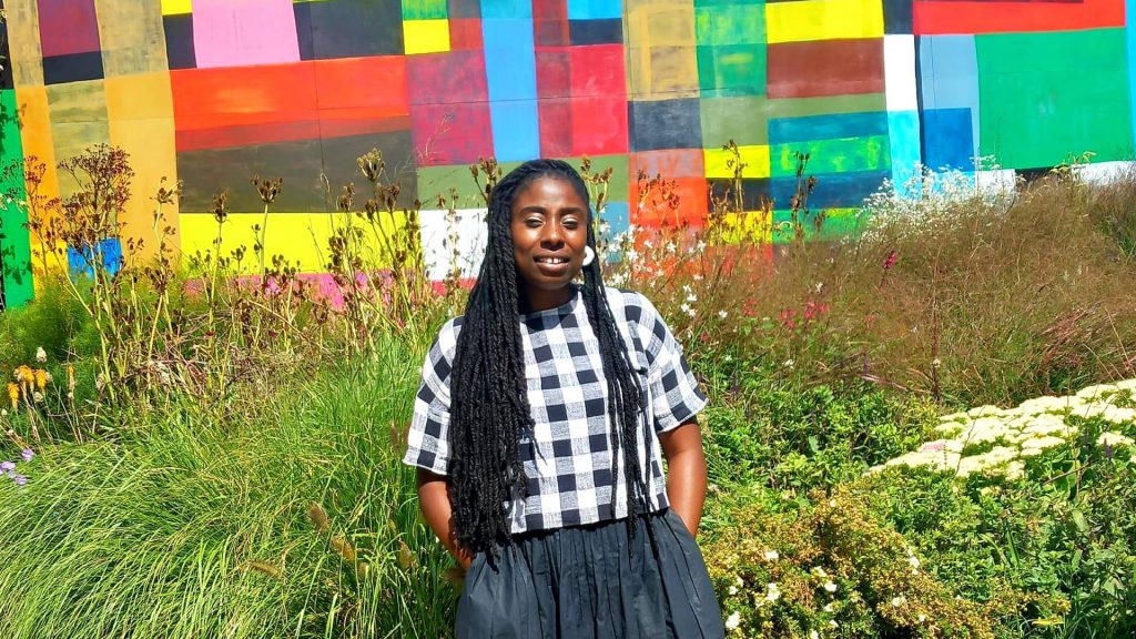 Kerri Akiwowo standing outside in front of a colourful painted wall and wild flowers.