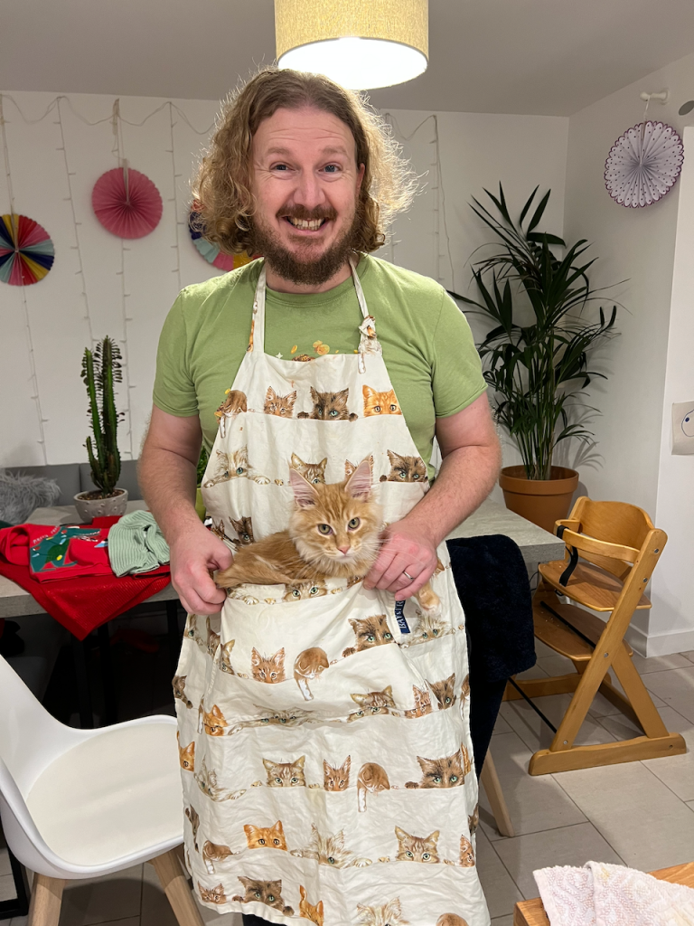 Oli Buckley wearing a cat pattern apron with a cat sitting in the apron pocket.