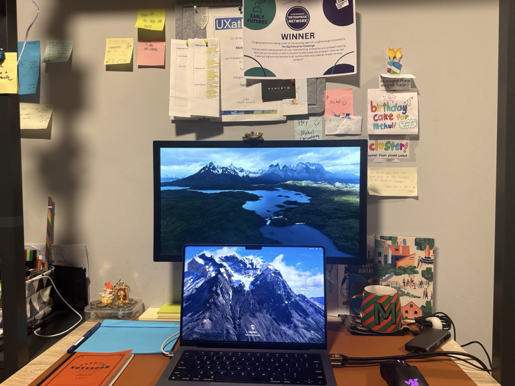 A desk with a laptop, monitor, mouse, mug, a notepad and pen on it. A variety of sticky notes and pieces of paper are on the pin board behind the desk