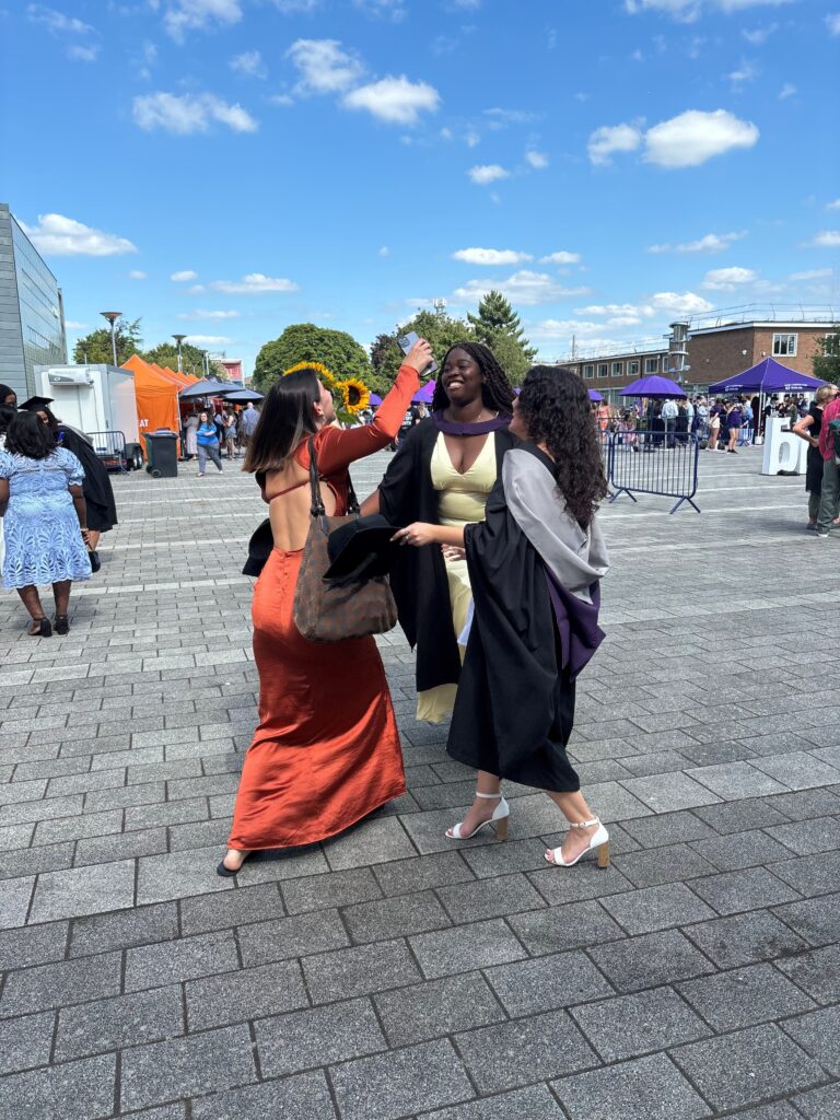 Lauryn in a cap and gown running and hugging 2 friends, 1 of whom is also in a cap nd gown. In the Shirley Pearce Square