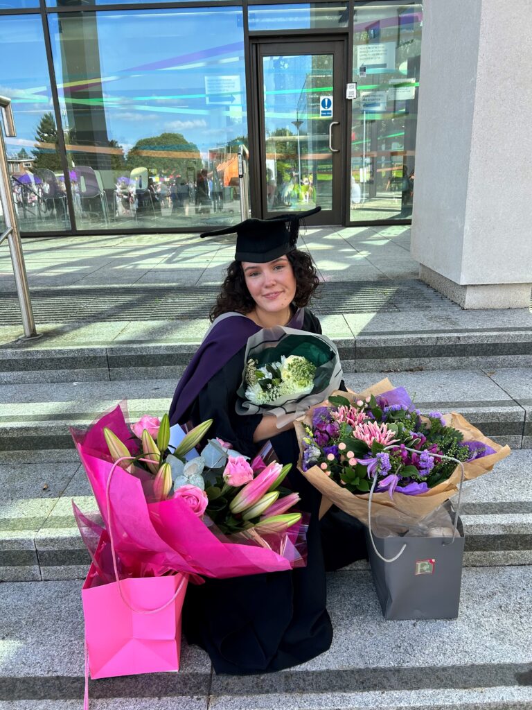 Lauryn crouching in a cap and gown with 3 bunches of flowers