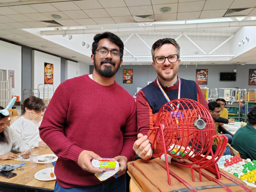 Two men standing with a bingo machine while groups of people play bingo in the background