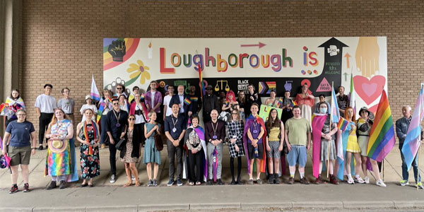 A group of staff and students standing in front of the 'Loughborough is all of us' mural on campus