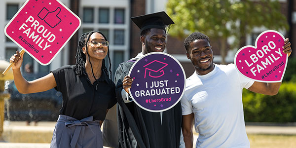 A graduating student in robes and hat with two supporters holding 'Proud family' and 'I just graduated' signs