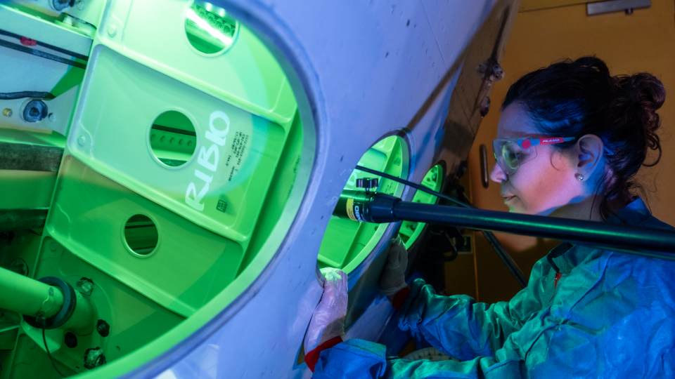 A person wearing a lab coat and goggles, looking into a machine.