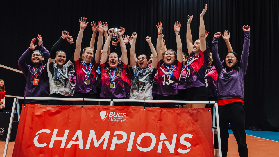 Volleyball team lift the championship trophy in front of a BUCS banner reading 'Champions'