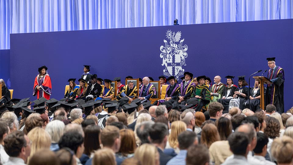 Chief Operating Officer Richard Taylor addresses students and guests from a podium while other staff sit on stage in academic gowns